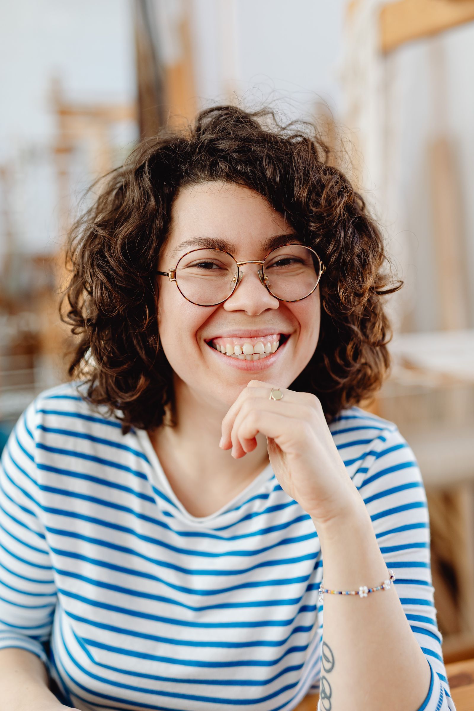 Portrait of a cheerful woman with curly hair and eyeglasses, wearing a striped shirt indoors.