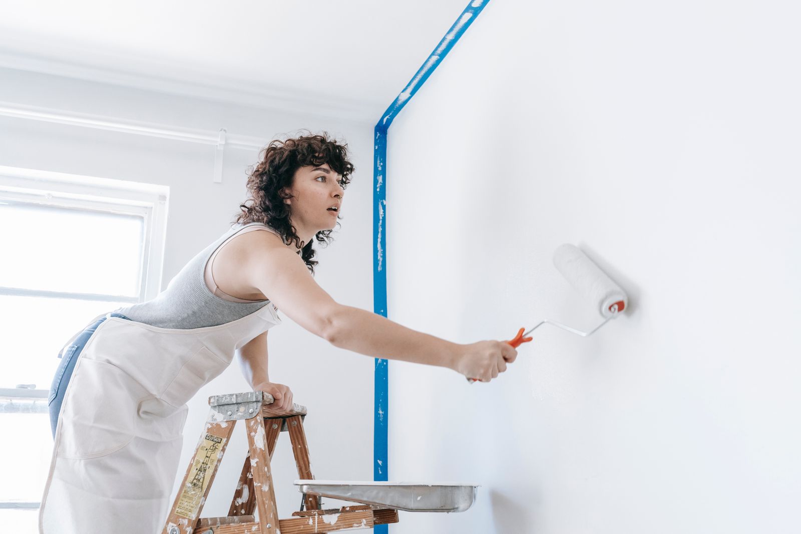A woman stands on a ladder painting a wall white, part of a DIY home renovation.